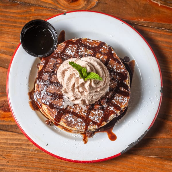 Chocolate chip pancakes with chocolate syrup, whipped cream, and a mint sprig on a white plate.