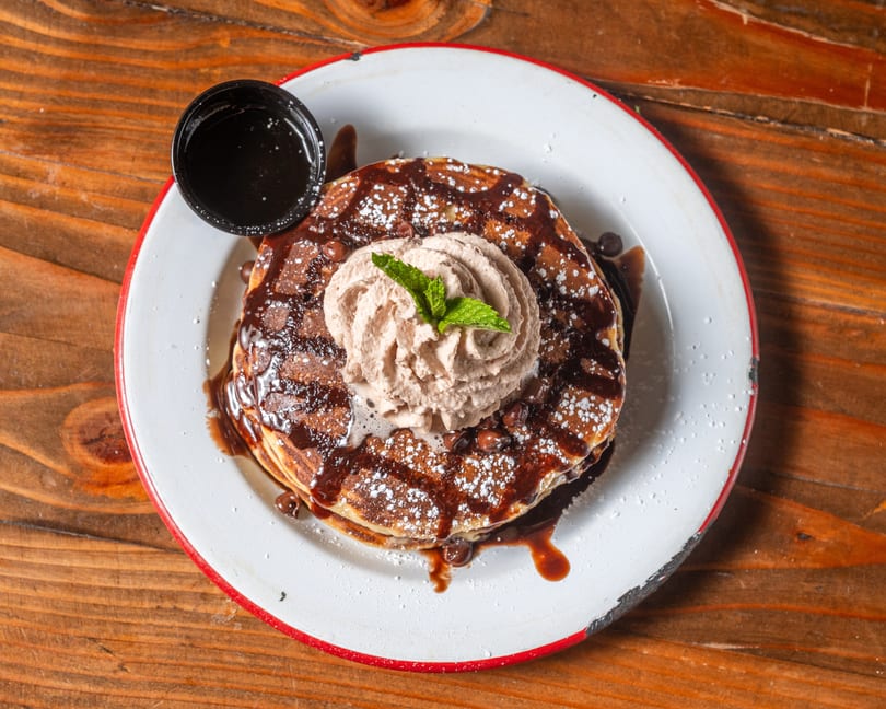 Chocolate chip pancakes with chocolate syrup, whipped cream, and a mint sprig on a white plate.