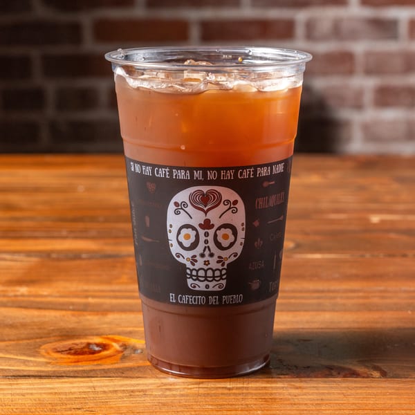 Iced coffee in a clear plastic cup with a decorative sugar skull label on a wooden table.