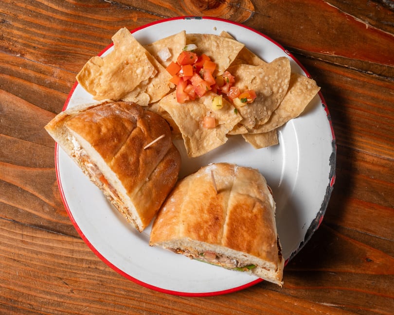 Two halves of a sandwich with chips and pico de gallo on a white plate with a red rim.