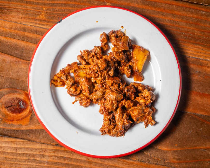 A white plate with a red rim holds a serving of saucy, chopped meat and pineapple chunks.