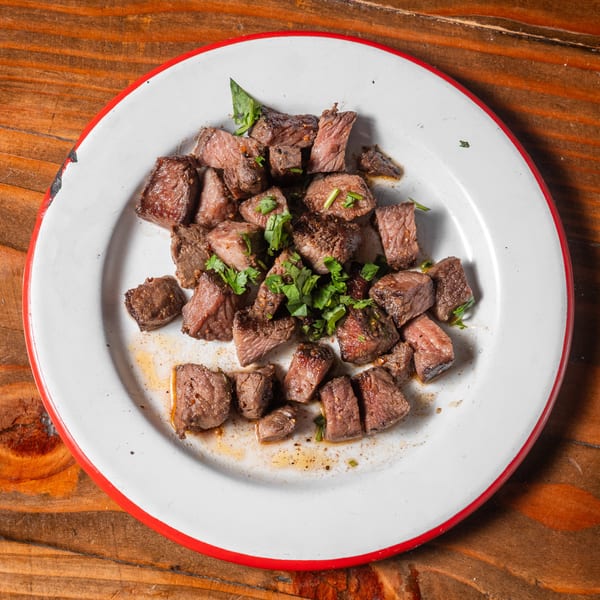 Cubed steak pieces with fresh herbs on a white plate with a red rim, on a wooden table.