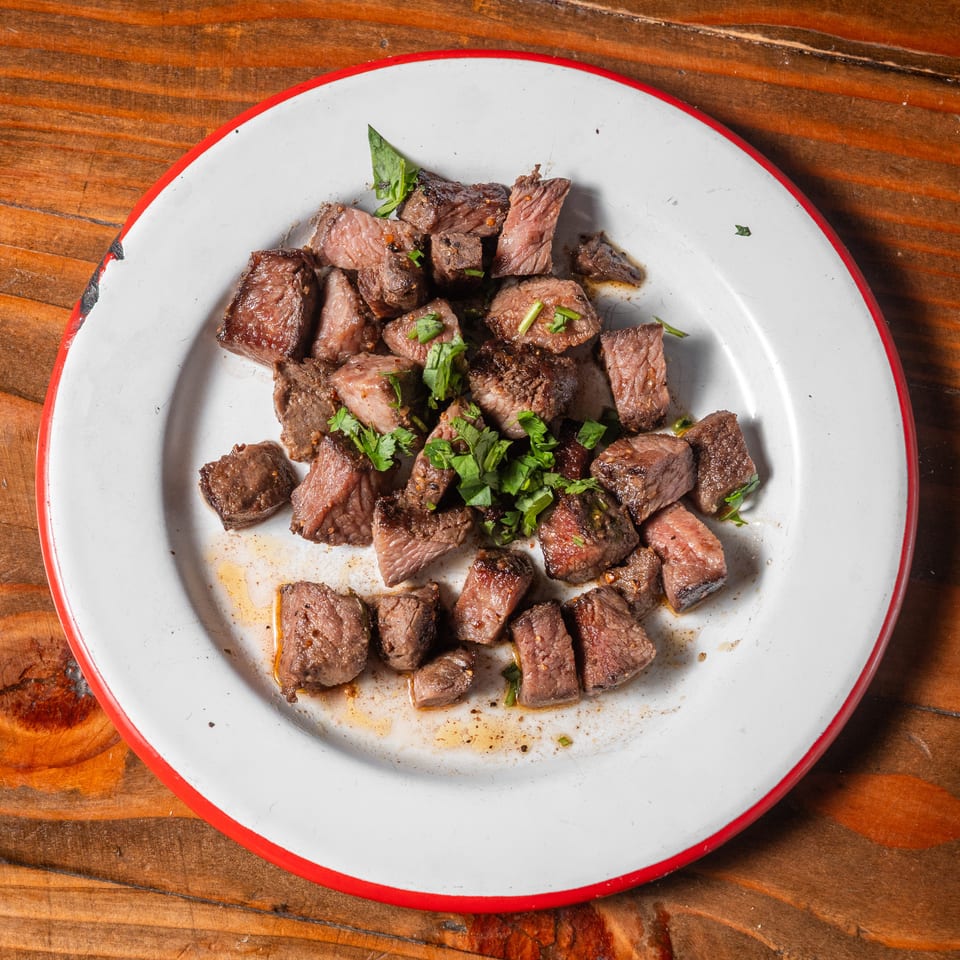 Cubed steak pieces with fresh herbs on a white plate with a red rim, on a wooden table.