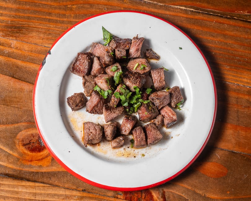 Cubed steak pieces with fresh herbs on a white plate with a red rim, on a wooden table.
