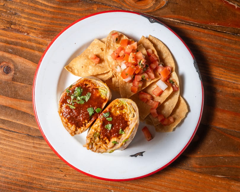 Two halves of a burrito topped with red sauce and cilantro, served with tortilla chips and pico de gallo.