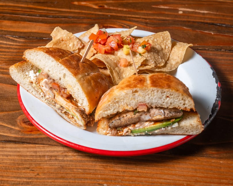 Two halves of a sandwich with chicken and avocado, served with tortilla chips and salsa on a white plate.