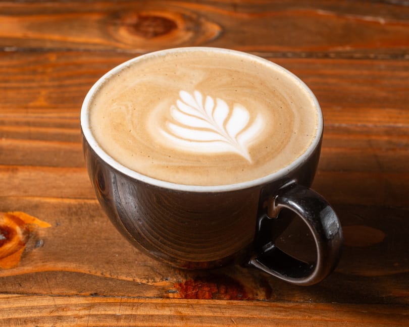 A dark ceramic mug of latte with intricate leaf-shaped latte art sits on a rustic wooden table.