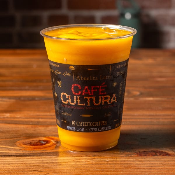 A Cafe Cultura mango drink in a clear plastic cup with a black label on a wooden table.