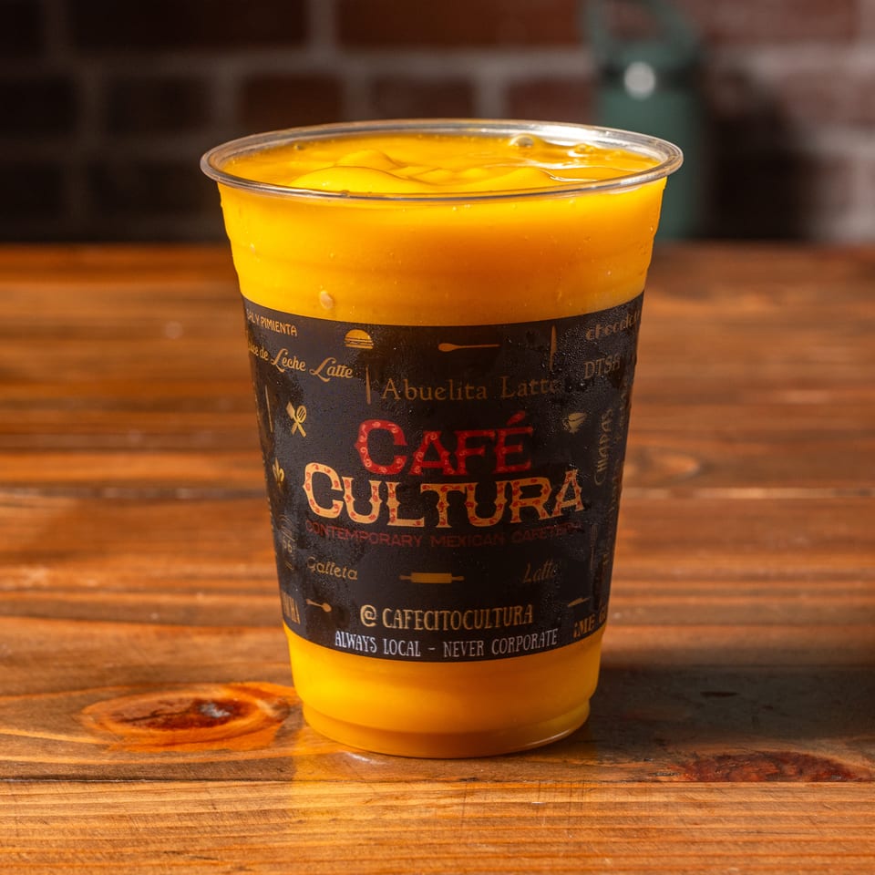 A Cafe Cultura mango drink in a clear plastic cup with a black label on a wooden table.