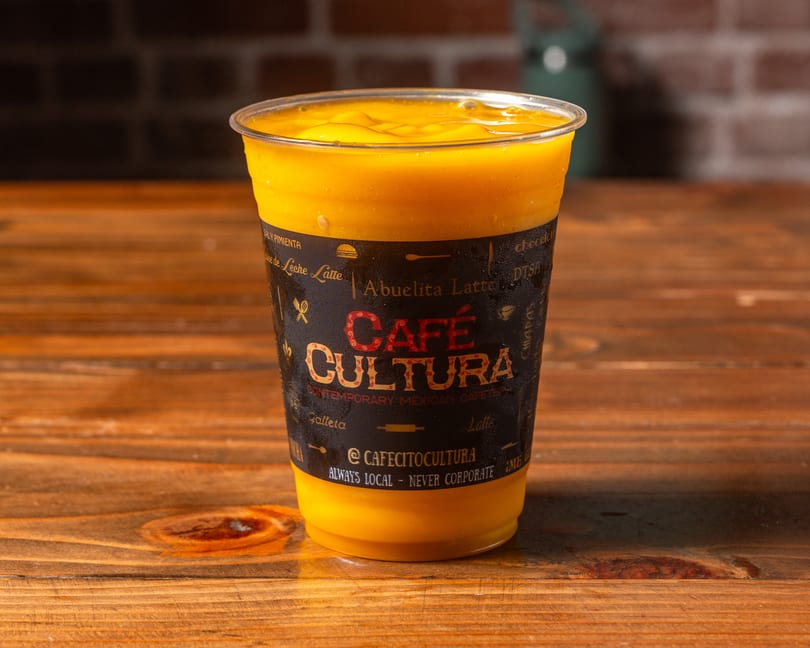 A Cafe Cultura mango drink in a clear plastic cup with a black label on a wooden table.