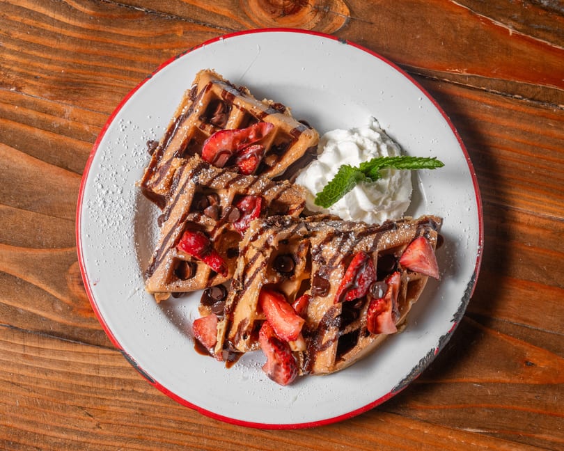 Two waffles topped with strawberries, chocolate chips, chocolate drizzle, powdered sugar, and whipped cream.