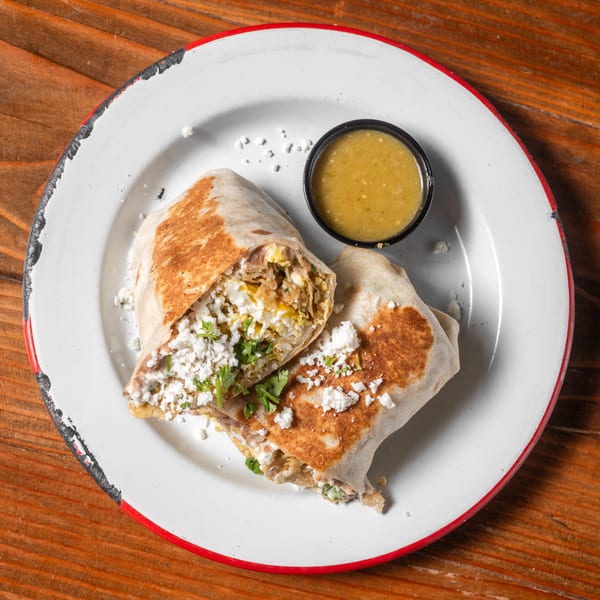 Two halves of a breakfast burrito with crumbled cheese and cilantro, served with green salsa.