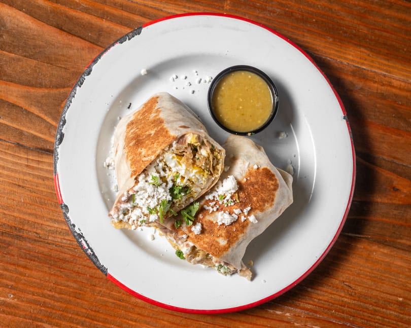 Two halves of a breakfast burrito with crumbled cheese and cilantro, served with green salsa.