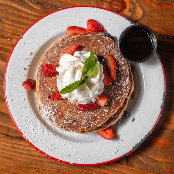 Stack of pancakes topped with whipped cream, strawberries, blueberries, and mint, with syrup on the side.