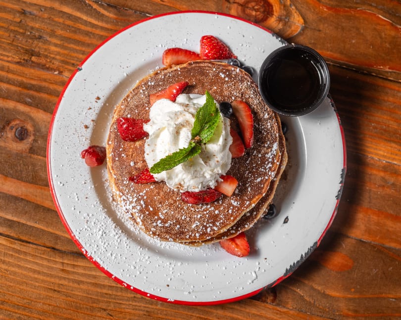 Stack of pancakes topped with whipped cream, strawberries, blueberries, and mint, with syrup on the side.