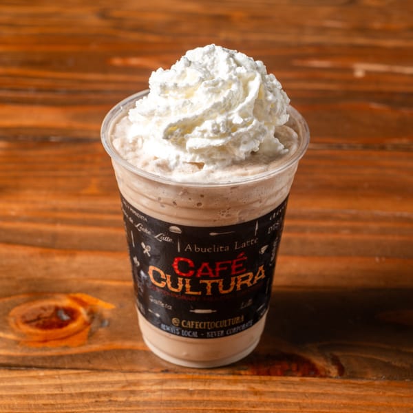 A Café Cultura Abuelita Latte, topped with whipped cream, sits on a rustic wooden table.