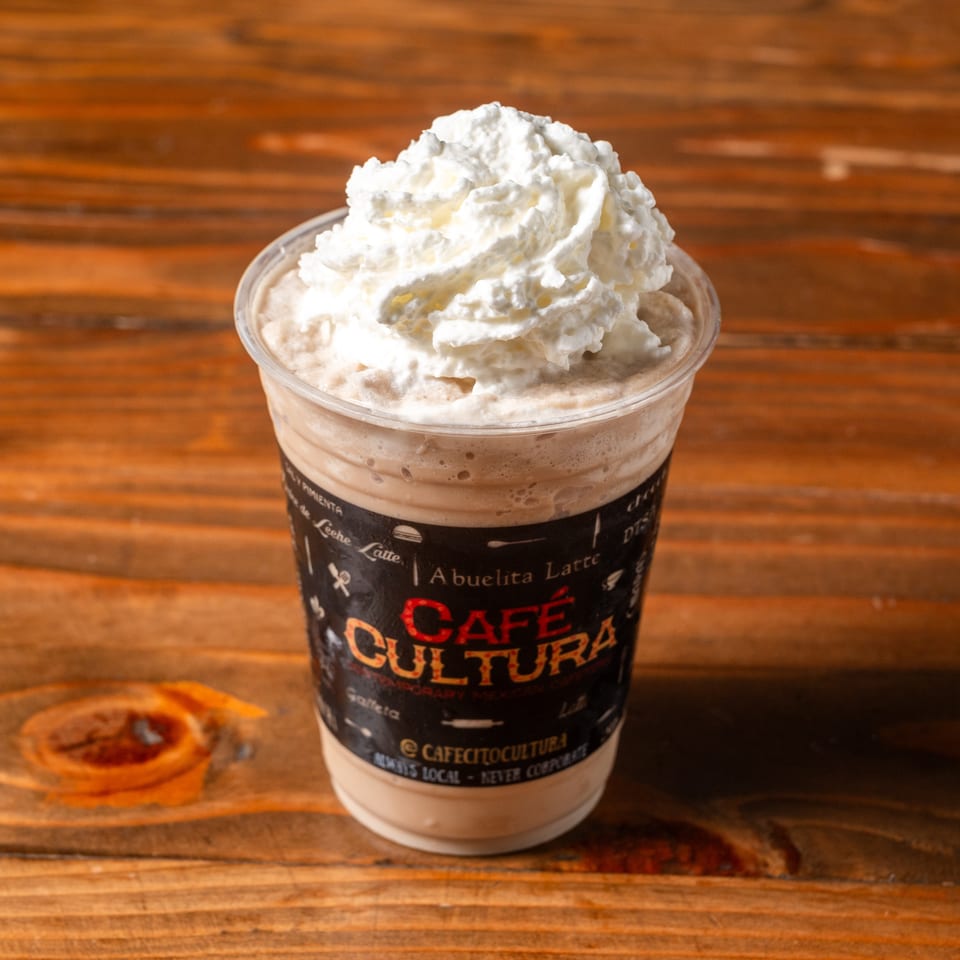 A Café Cultura Abuelita Latte, topped with whipped cream, sits on a rustic wooden table.