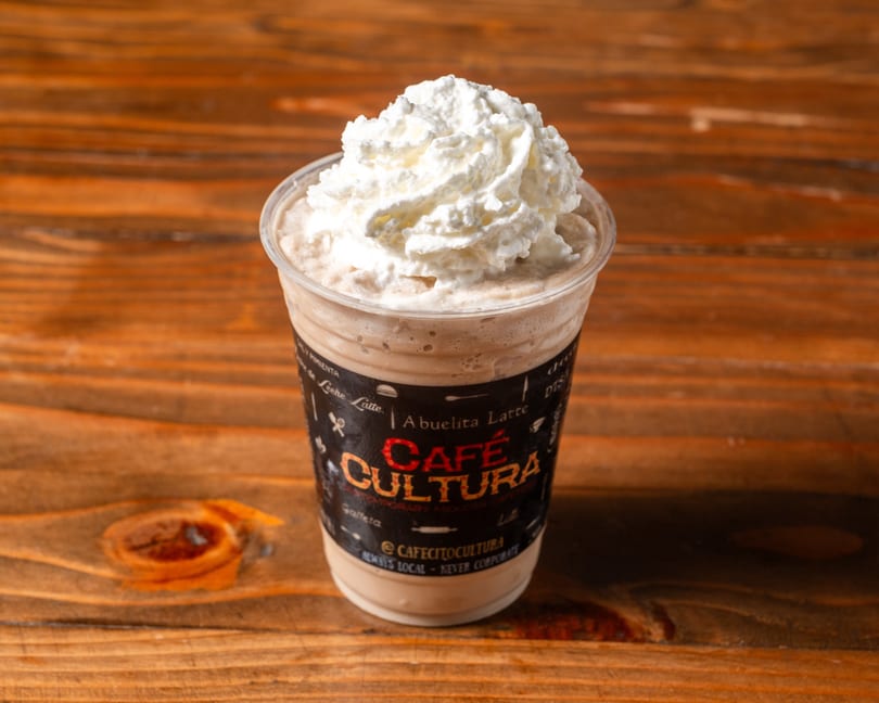 A Café Cultura Abuelita Latte, topped with whipped cream, sits on a rustic wooden table.