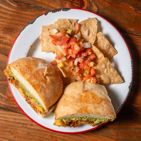 Two torta halves with guacamole and meat, served with tortilla chips and fresh salsa.