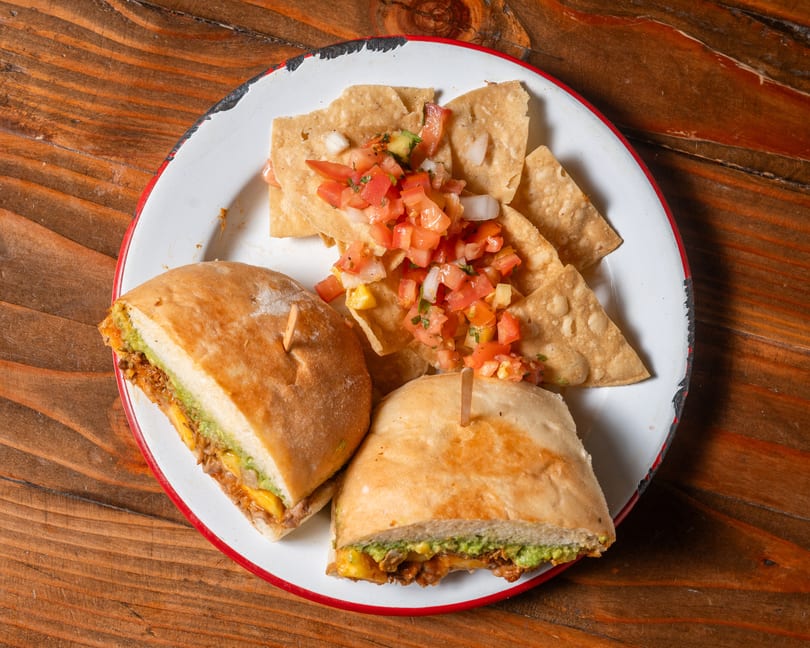 Two torta halves with guacamole and meat, served with tortilla chips and fresh salsa.