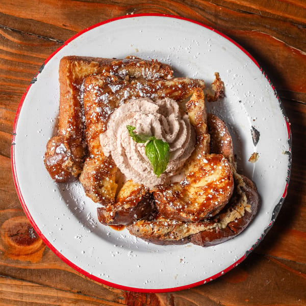 French toast with powdered sugar, syrup, whipped cream, and a mint leaf on a white plate.
