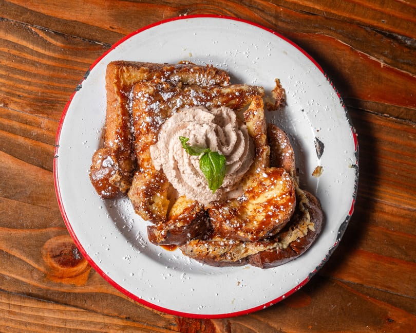 French toast with powdered sugar, syrup, whipped cream, and a mint leaf on a white plate.