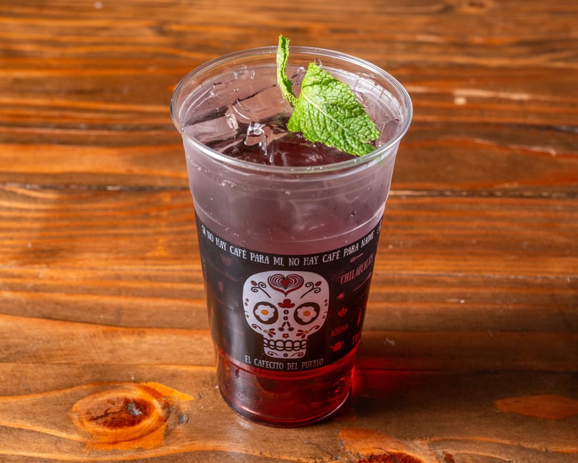 A vibrant red drink with ice and mint in a clear cup with a sugar skull design.
