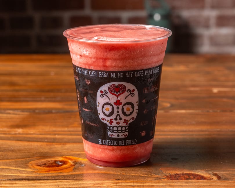 A vibrant red smoothie in a clear cup with a decorative black sleeve featuring a sugar skull design.