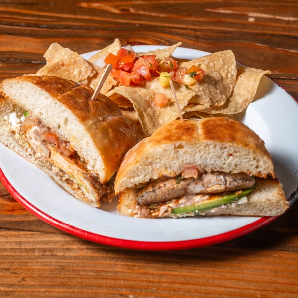 Two halves of a sandwich with chicken and avocado, served with tortilla chips and salsa on a white plate.