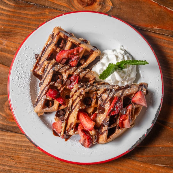 Two waffles topped with strawberries, chocolate chips, syrup, powdered sugar, and whipped cream with mint.