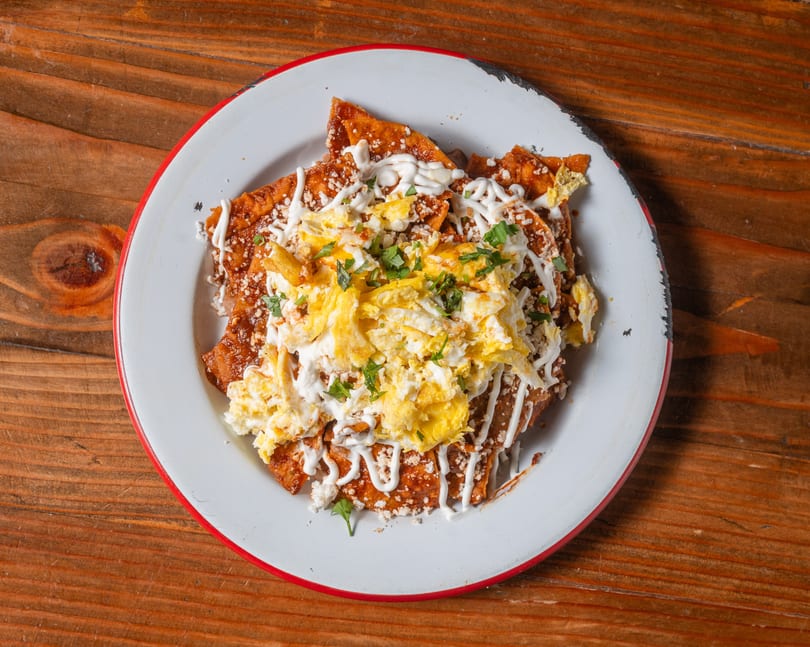 Chilaquiles with red sauce, scrambled eggs, sour cream, cheese, and cilantro on a white plate.